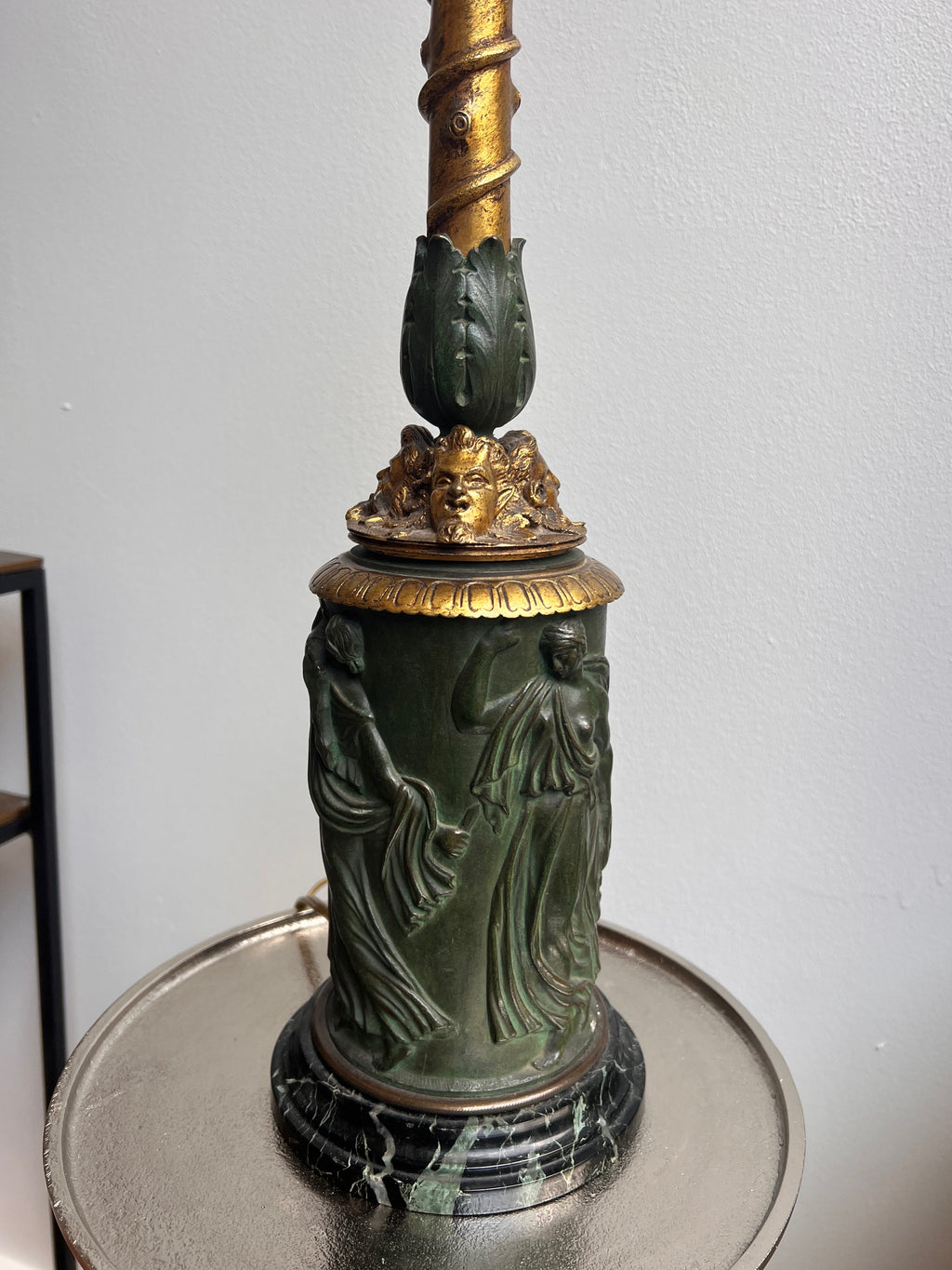 An ornate antique Neoclassical Revival table lamp with a dark green carved column featuring classical female figures, a gold-gilt decorative collar, and a matching gold-toned spiral stem, set on a round metallic tray against a plain light gray wall.