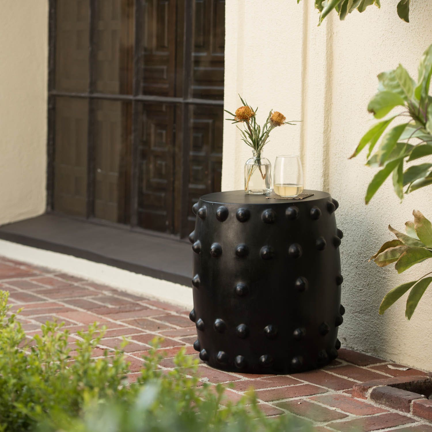 A black cylindrical garden stool with a matte finish and raised circular studs around the sides, styled in a luxury backyard setting.