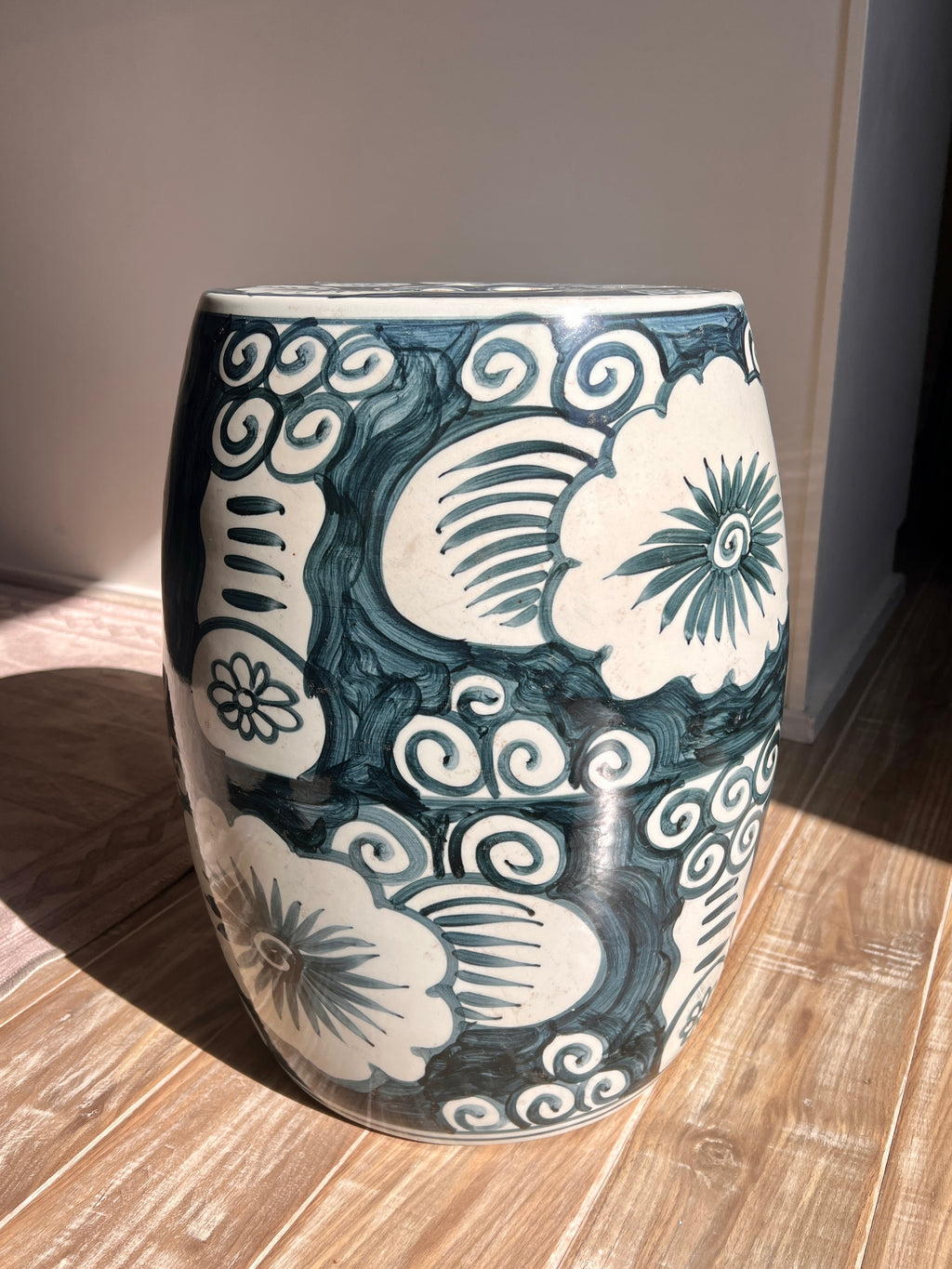 A round ceramic stool with a dark teal and cream floral cloud pattern, photographed on a wooden floor in natural sunlight.