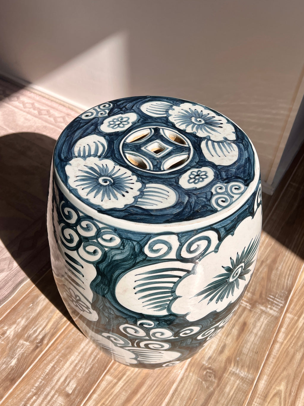 A round ceramic garden stool with a blue and white floral pattern, featuring a cutout coin-like design on the top, set on a wooden floor.