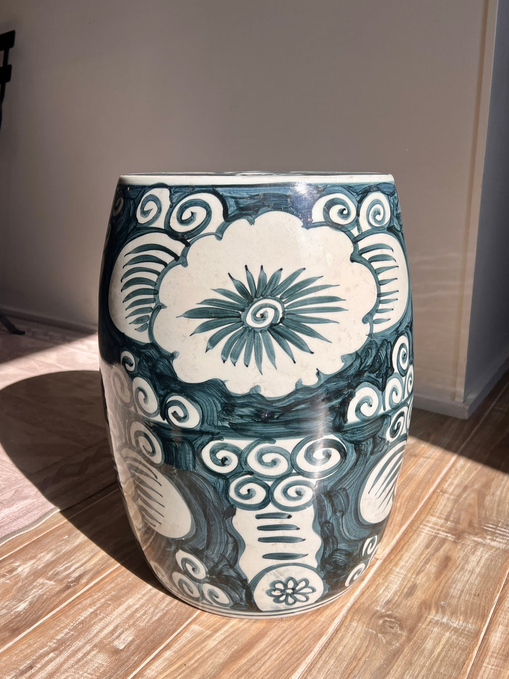 A cylindrical ceramic garden stool with a glossy dark teal and off-white hand-painted floral and scroll pattern, standing on a light wood floor in a room.
