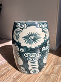 A cylindrical ceramic garden stool with a glossy dark teal and off-white hand-painted floral and scroll pattern, standing on a light wood floor in a room.