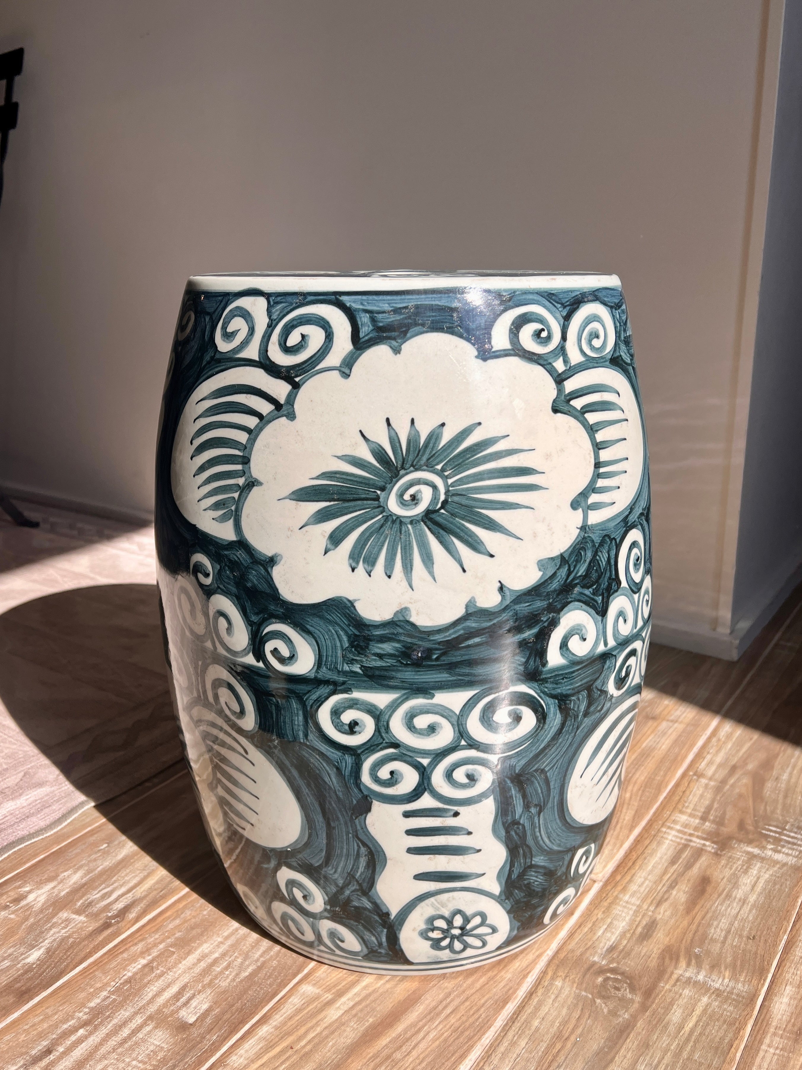 A cylindrical ceramic garden stool with a glossy dark teal and off-white hand-painted floral and scroll pattern, standing on a light wood floor in a room.
