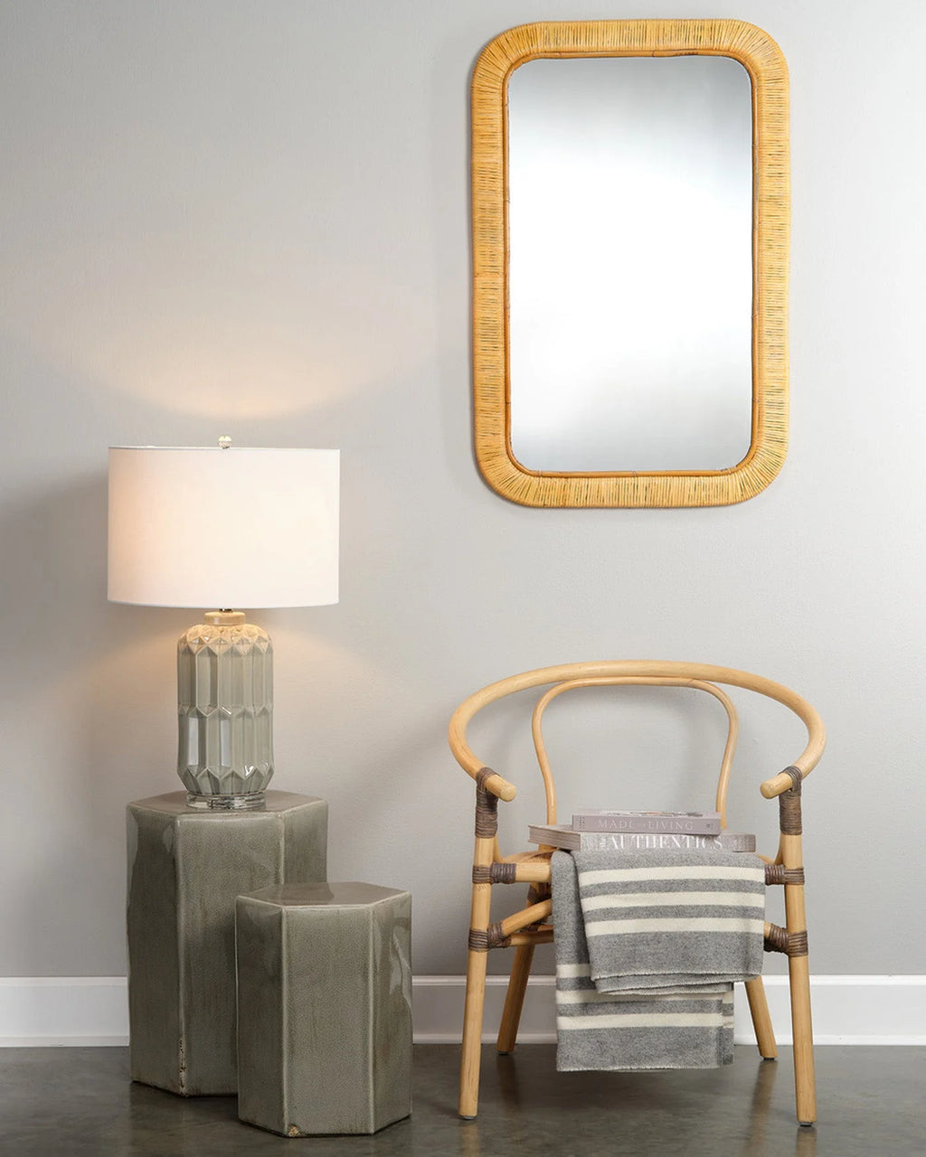 A rectangular wall mirror with a natural woven rattan frame hangs on a light gray wall above a wooden chair with a striped throw blanket. To the left, a table lamp with a white drum shade and a faceted gray glass base sits on two stacked gray side tables.