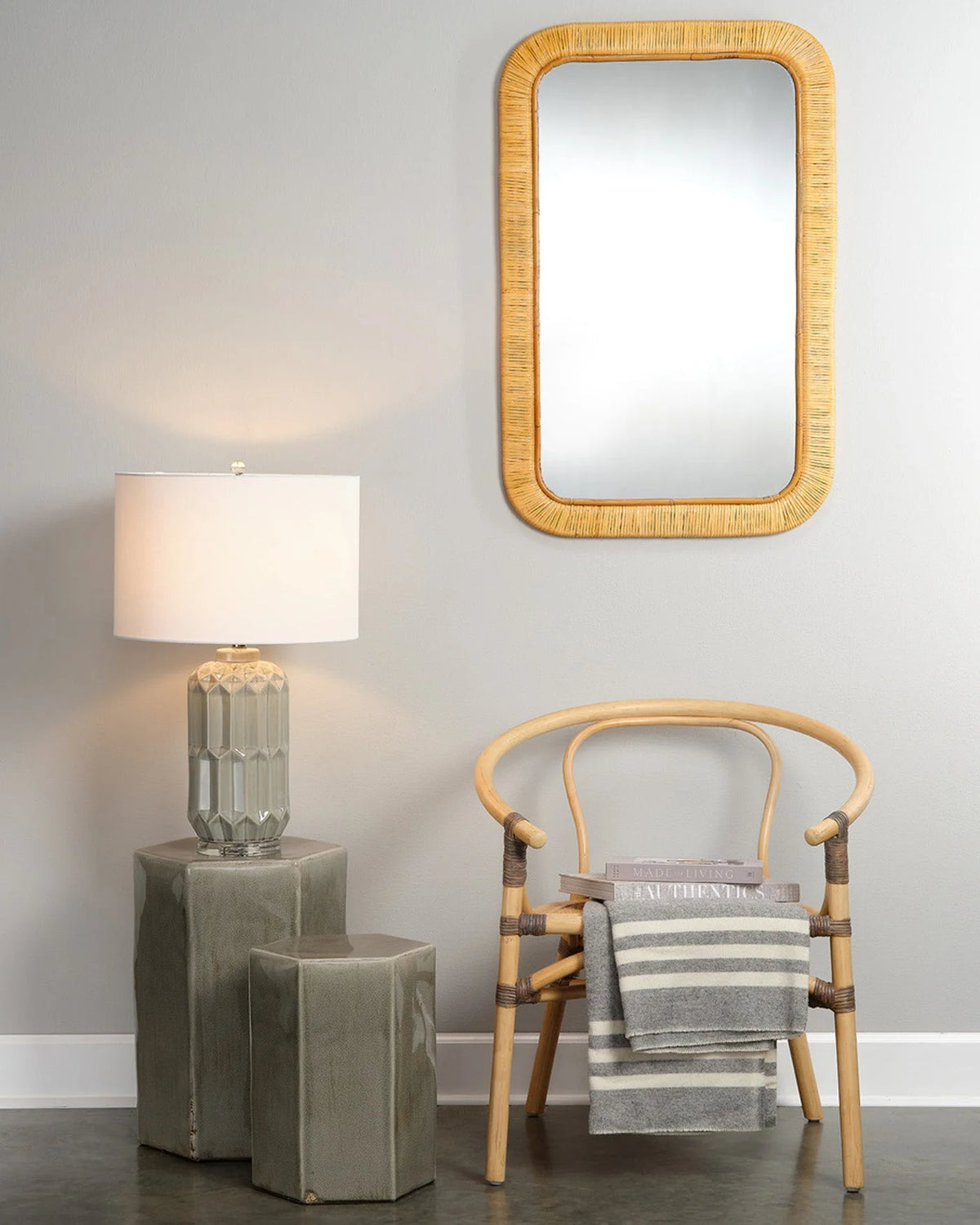 A rectangular wall mirror with a natural woven rattan frame hangs on a light gray wall above a wooden chair with a striped throw blanket. To the left, a table lamp with a white drum shade and a faceted gray glass base sits on two stacked gray side tables.