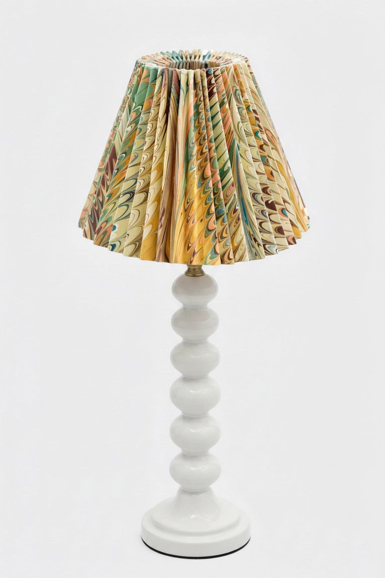 A table lamp with a pleated conical shade featuring a multicolor abstract pattern in yellow, green, brown, and beige, mounted on a glossy white turned-bead lamp base against a light gray background.