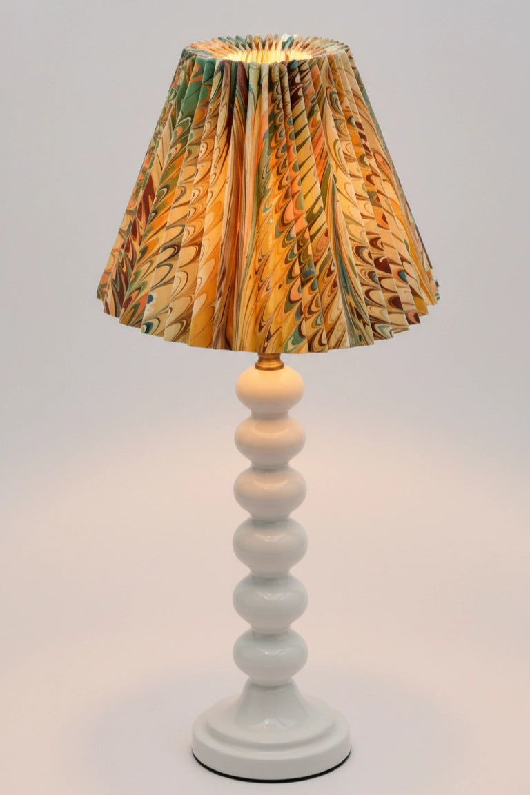 A table lamp with a white turned wood bead spindle base and a pleated fabric shade featuring a marbled pattern of gold, brown, and green.