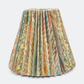 A pleated table lampshade featuring a marbled paper pattern with swirls of beige, green, orange, and brown, set against a plain light gray background.