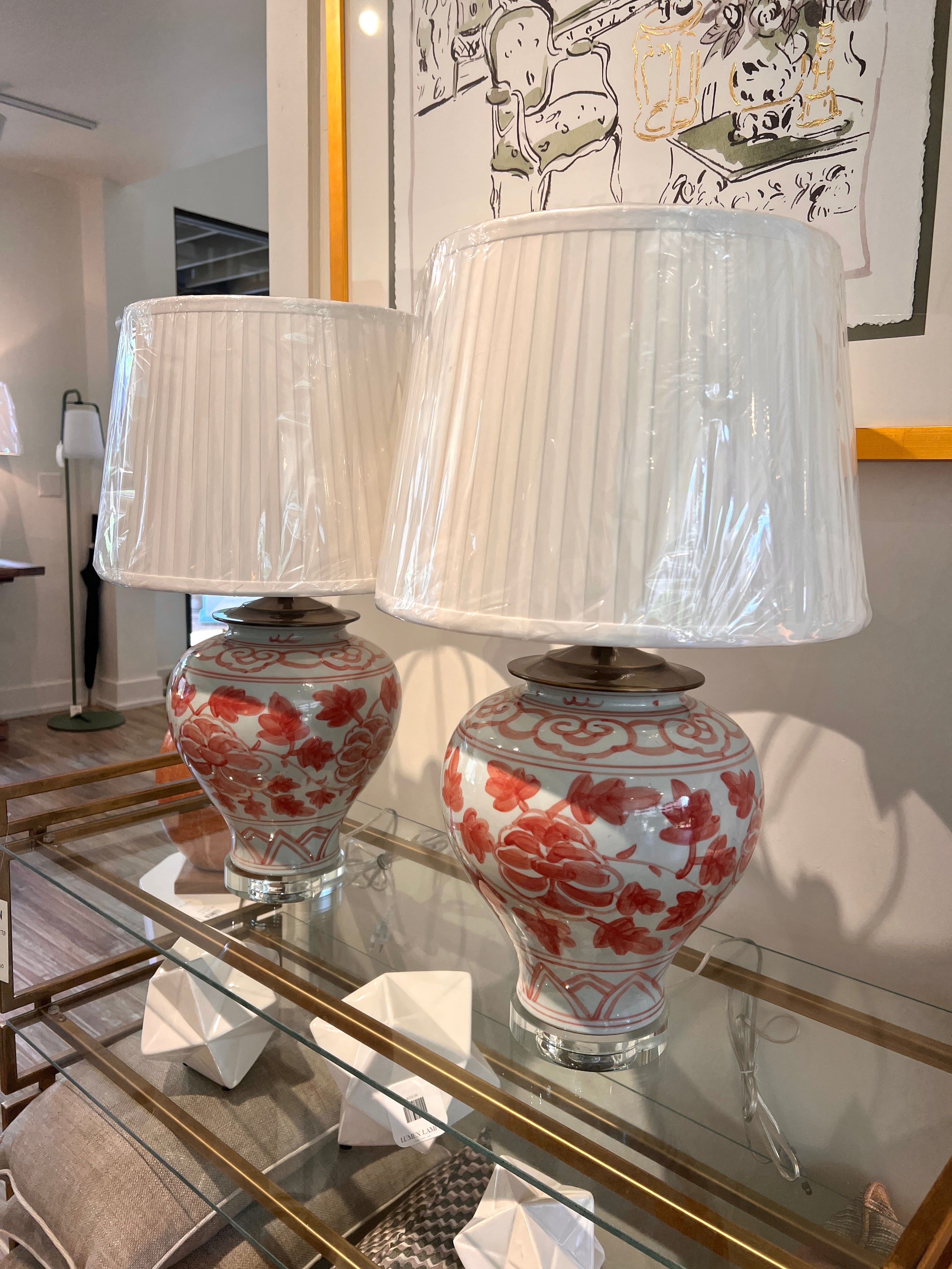 Two matching table lamps with white pleated drum shades and glossy white ceramic bases decorated with pink floral Asian motifs, displayed on a glass-topped console table in an indoor room.