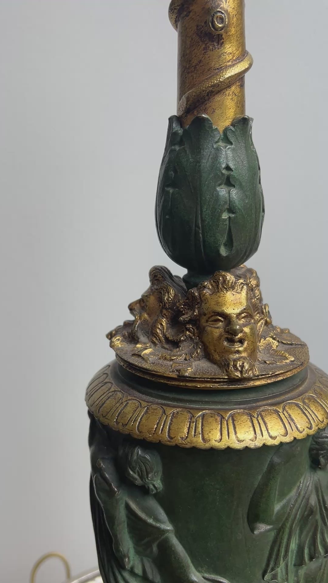 An ornate antique Neoclassical Revival table lamp with a dark green carved column featuring classical female figures, a gold-gilt decorative collar, and a matching gold-toned spiral stem, set on a round metallic tray against a plain light gray wall.