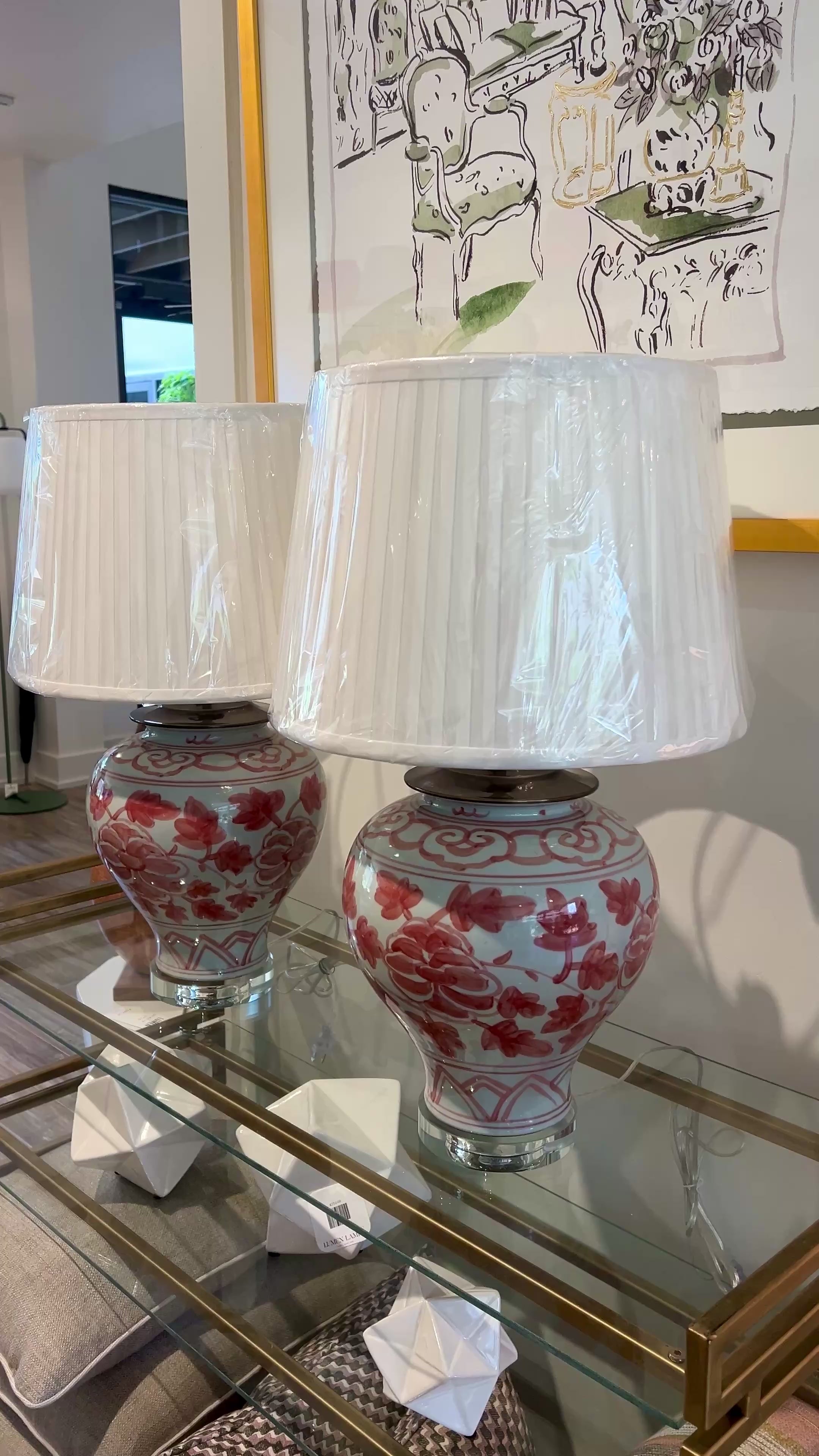 Two matching table lamps with white pleated drum shades and glossy white ceramic bases decorated with pink floral Asian motifs, displayed on a glass-topped console table in an indoor room.