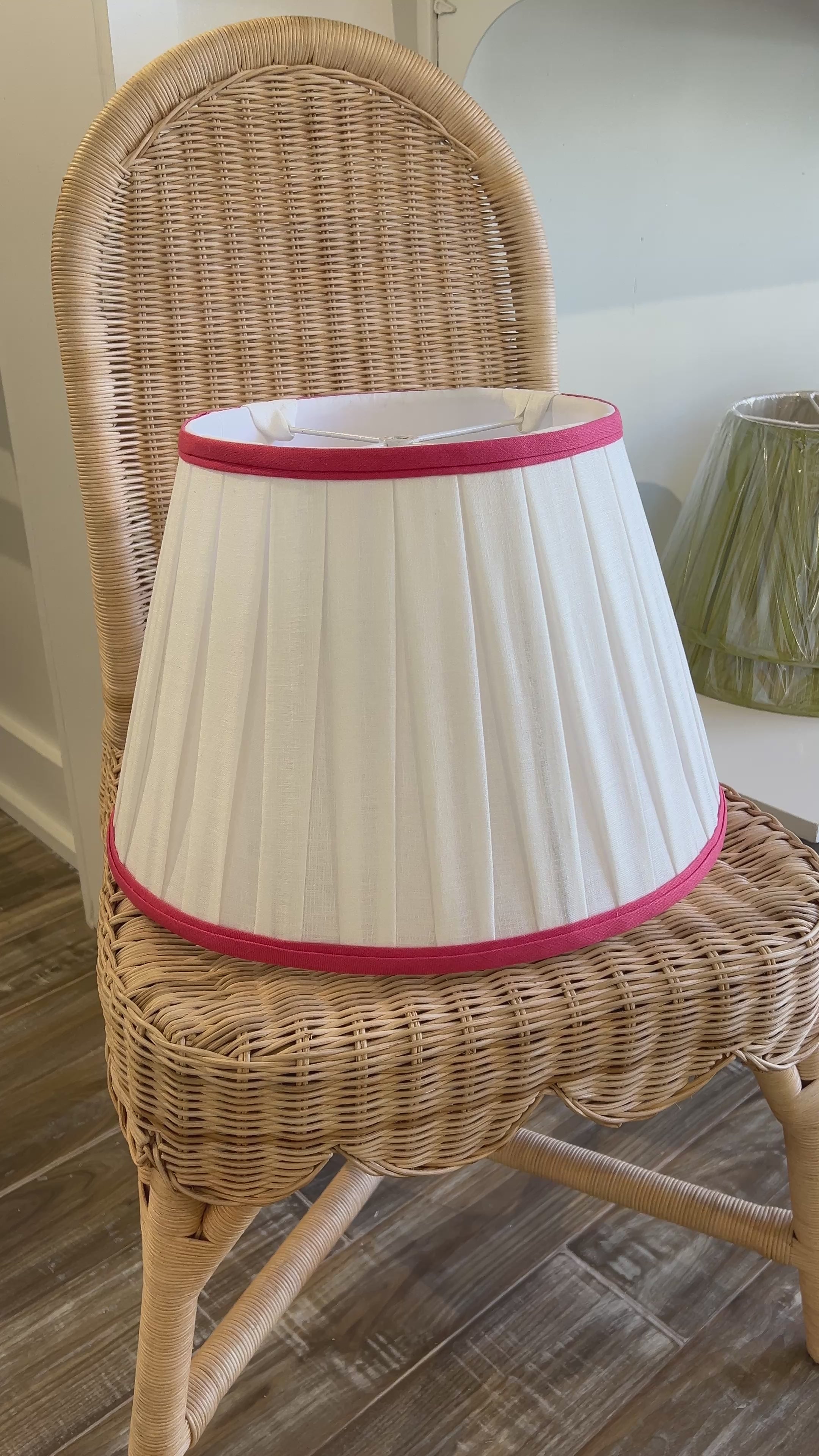 A natural wicker dining chair with a pleated white fabric lampshade and pink trim, set on a wooden floor in a home interior.
