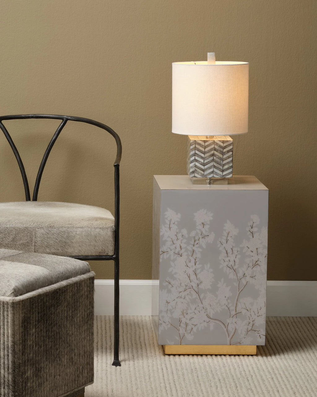 A modern table lamp with a white drum shade and a gray chevron-patterned base sits on a square gray side table with a floral branch print and a gold base, styled in a living room next to a gray chair.