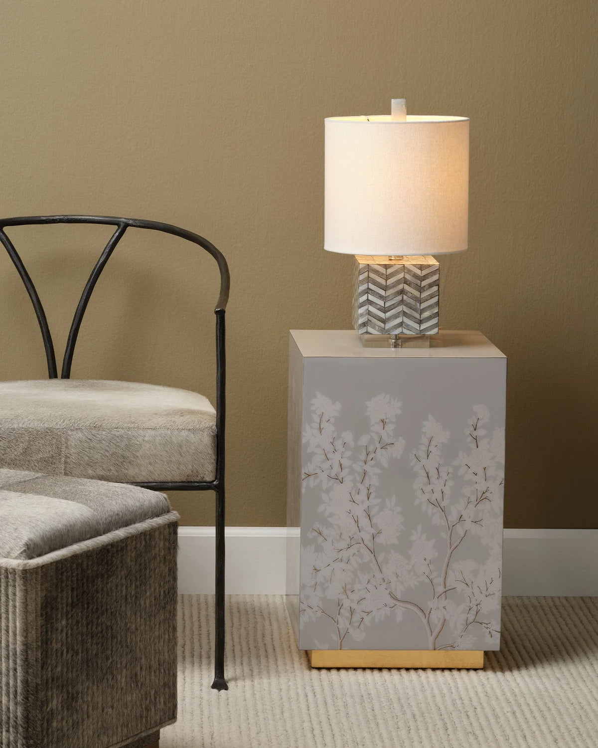 A modern table lamp with a white drum shade and a gray chevron-patterned base sits on a square gray side table with a floral branch print and a gold base, styled in a living room next to a gray chair.