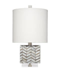 A table lamp with a large white drum shade, a square base made of inlaid mother-of-pearl chevron pattern, and a clear acrylic foot.