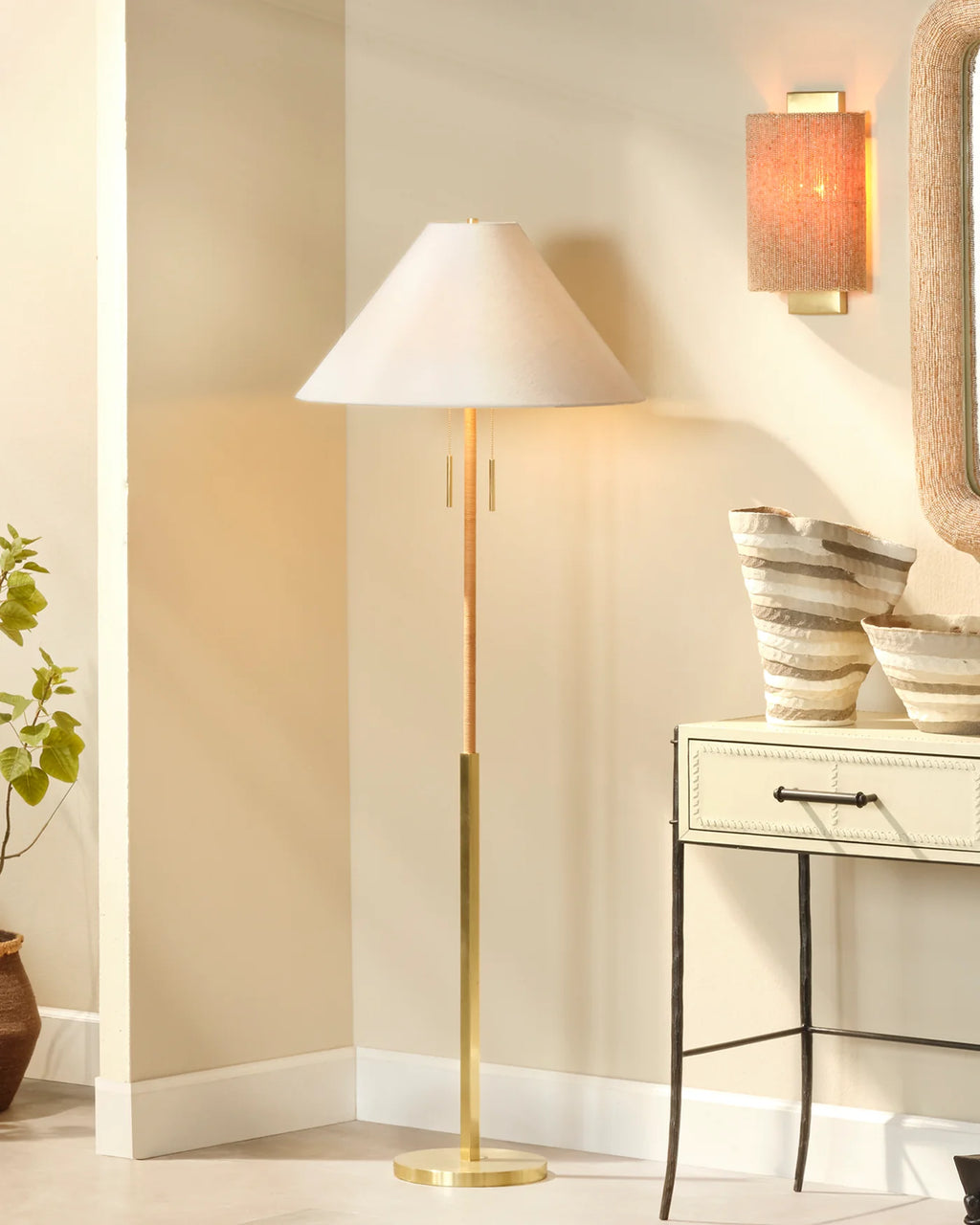 A modern floor lamp with a tapered off-white shade and a brushed gold metal stand and base, styled in a warm, beige-toned room next to a console table and a wall sconce.