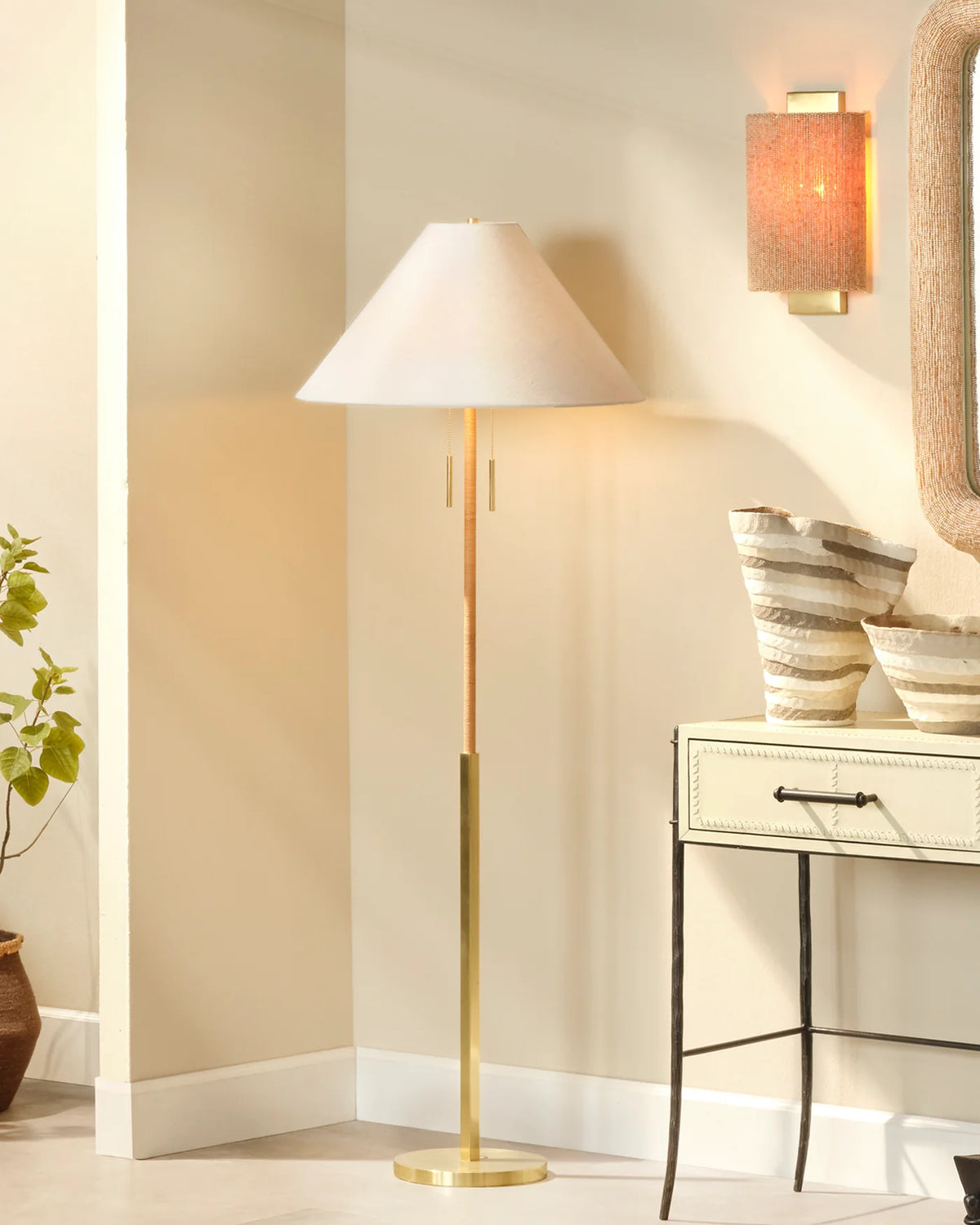 A modern floor lamp with a tapered off-white shade and a brushed gold metal stand and base, styled in a warm, beige-toned room next to a console table and a wall sconce.