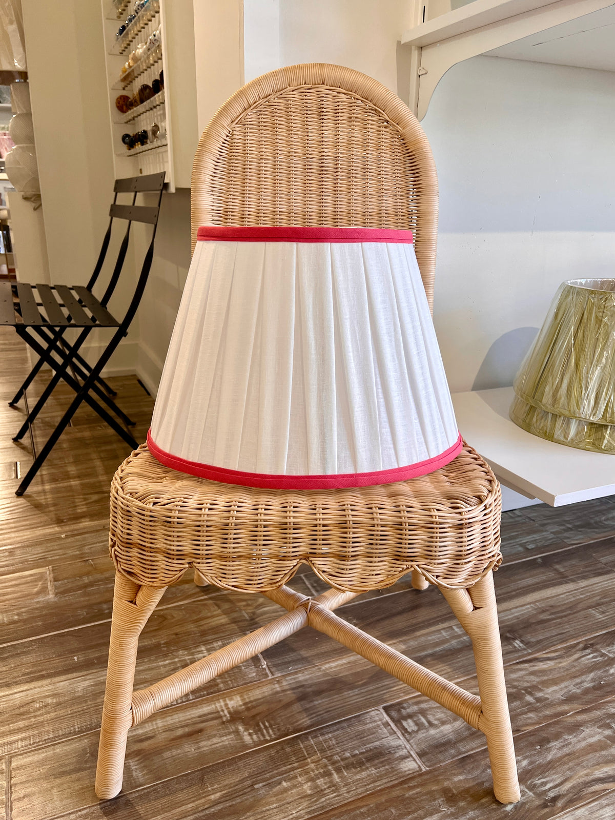 A natural wicker dining chair with a pleated white fabric lampshade and pink trim, set on a wooden floor in a home interior.